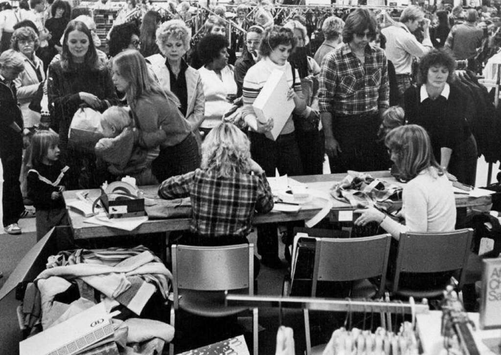 A crowded store with people lined up. Women are sitting at a table near piles of clothes, assisting customers. Shoppers, including children, are engaged in purchasing or interacting with staff. Clothing racks are visible in the background. Black and white image.