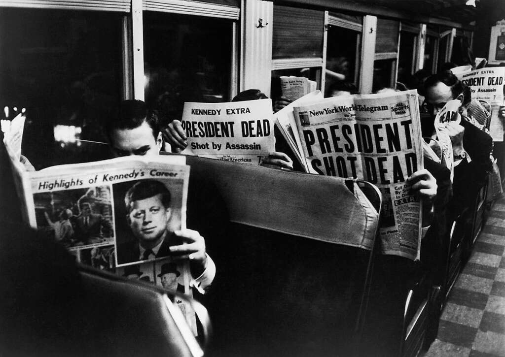 Passengers sitting in a train meticulously read newspapers with headlines announcing the death of the president. The photo captures a historical moment of shared solemnity and shock.