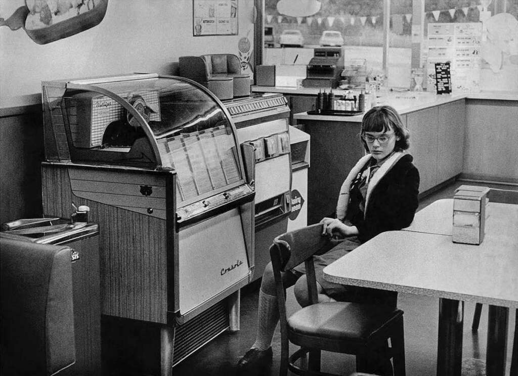 A woman sits alone in a vintage diner next to a jukebox. The room contains a few chairs and tables, and the walls are decorated with signs. Large windows in the background show a view of parked cars outside. The scene is in black and white.