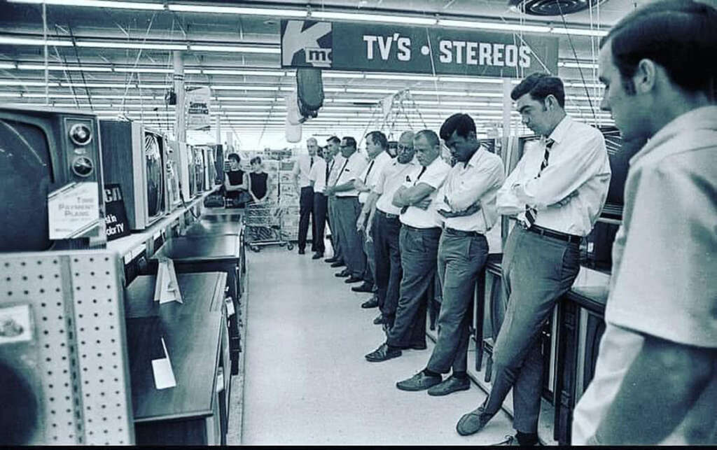 A group of men in a store stands intently watching several TV screens. They are dressed in shirts and ties, some with arms crossed. The aisle is lined with TV sets and other electronics. A sign above reads "TV's - Stereos.