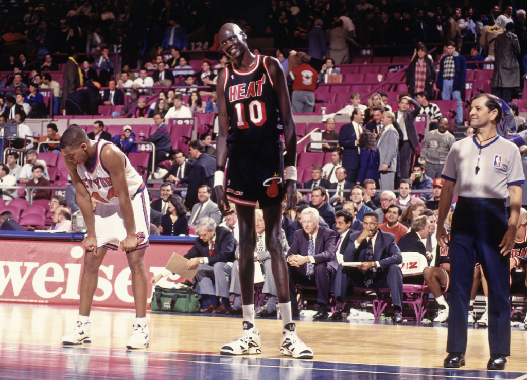 A basketball game with a player from the Miami Heat and another from the New York Knicks standing on the court. The Heat player, wearing jersey number 10, is noticeably taller. An NBA referee stands beside them. The stands are filled with spectators.