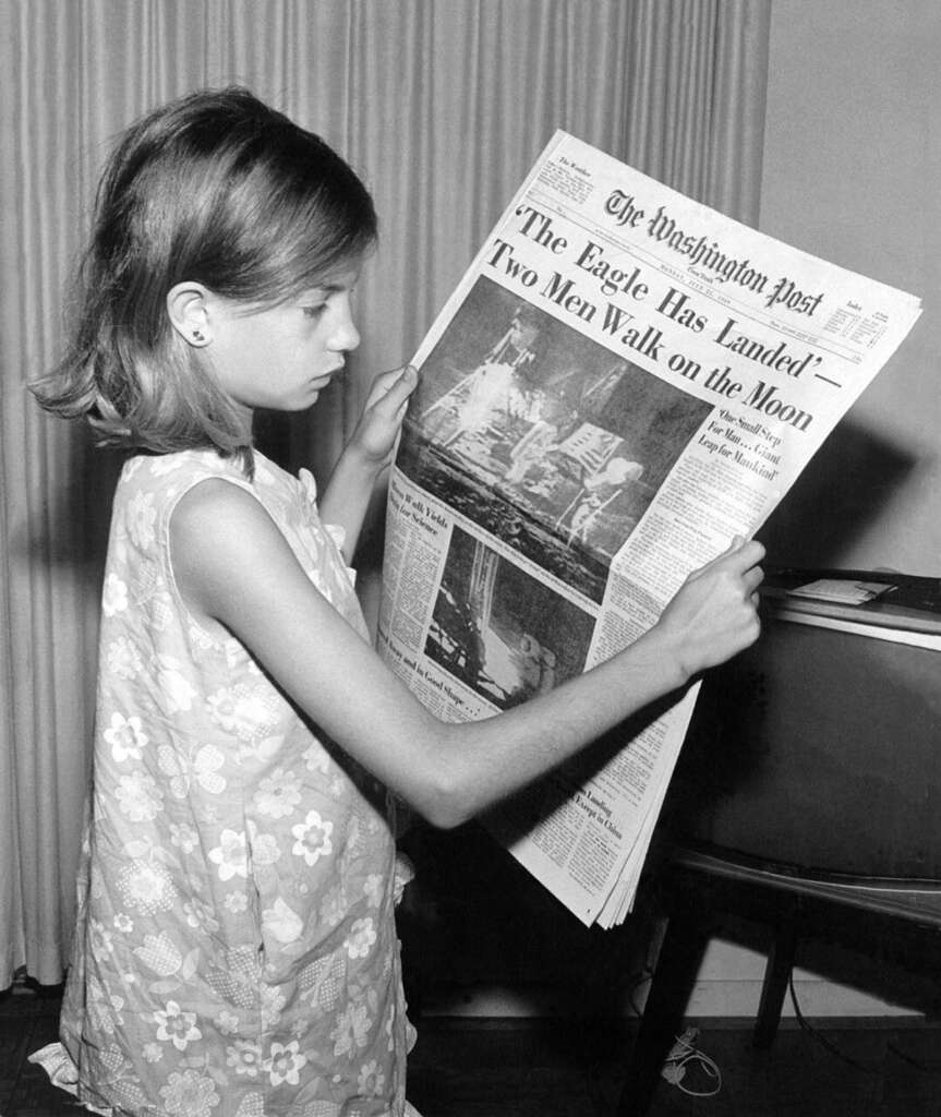 A young girl in a floral dress reads a vintage newspaper with the headline "'The Eagle Has Landed'—Two Men Walk on Moon." The black and white photo shows her standing next to a TV in a room with curtains.