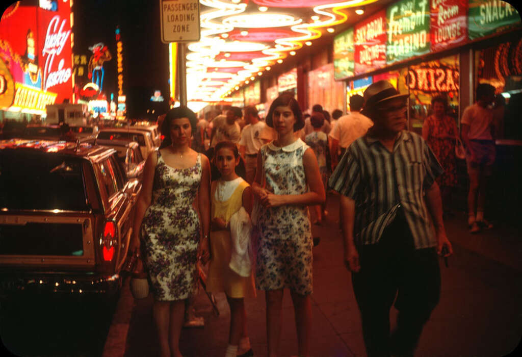 A group of people walk along a brightly lit street at night, filled with colorful neon signs and bustling with activity. A woman and two girls in patterned dresses stand next to a car, while others stroll past in casual summer attire.