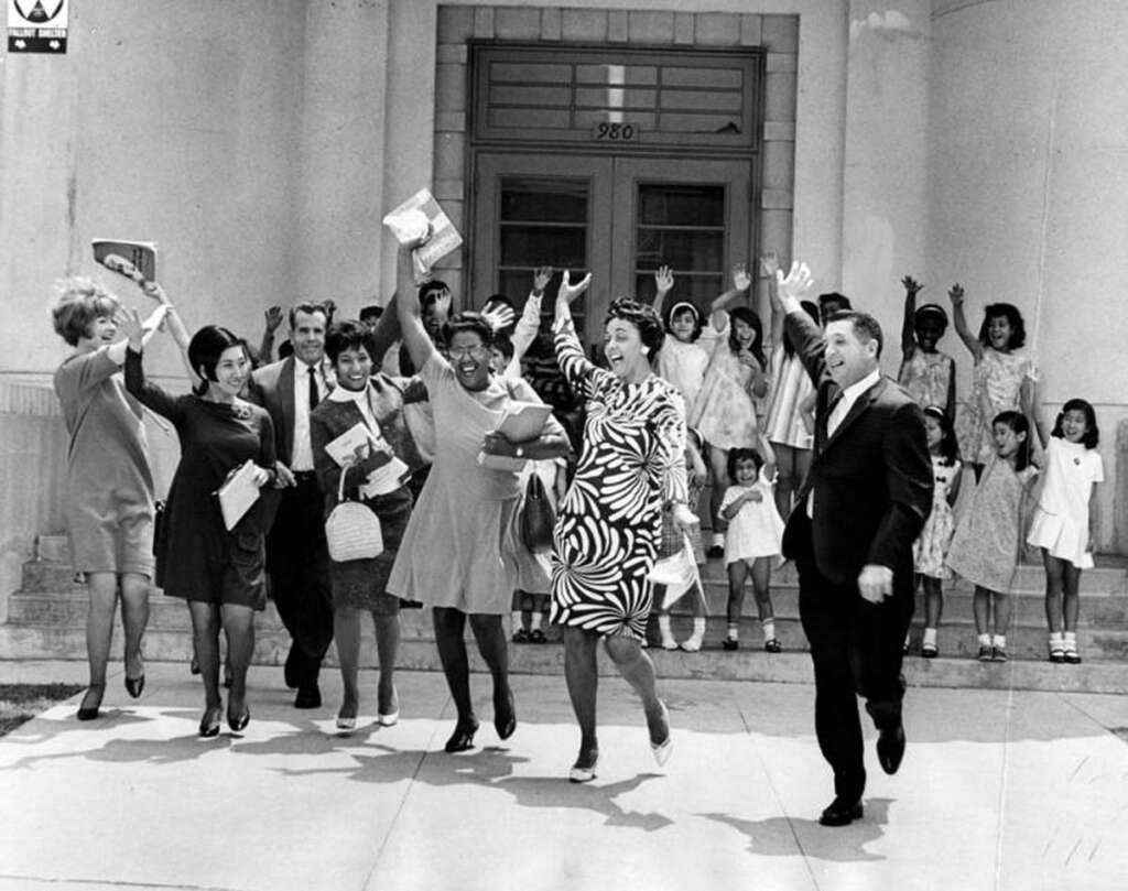 A group of people cheerfully raise their arms while exiting a building, carrying books and papers. They're dressed in mid-20th century attire, with both men and women in the group. Children stand in the background near the entrance.