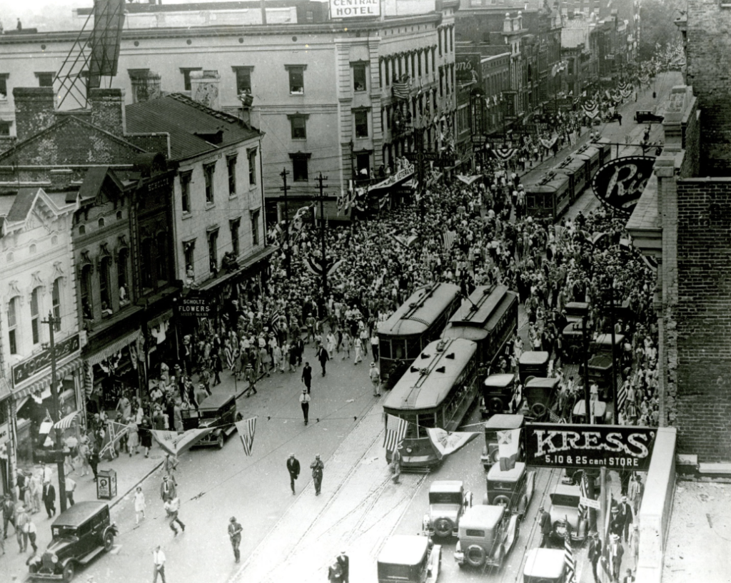 A bustling street scene from the early 20th century shows a large crowd gathered for a parade. Trolley cars run down the center of the street, surrounded by old buildings with a store sign reading "KRESS" visible in the foreground.