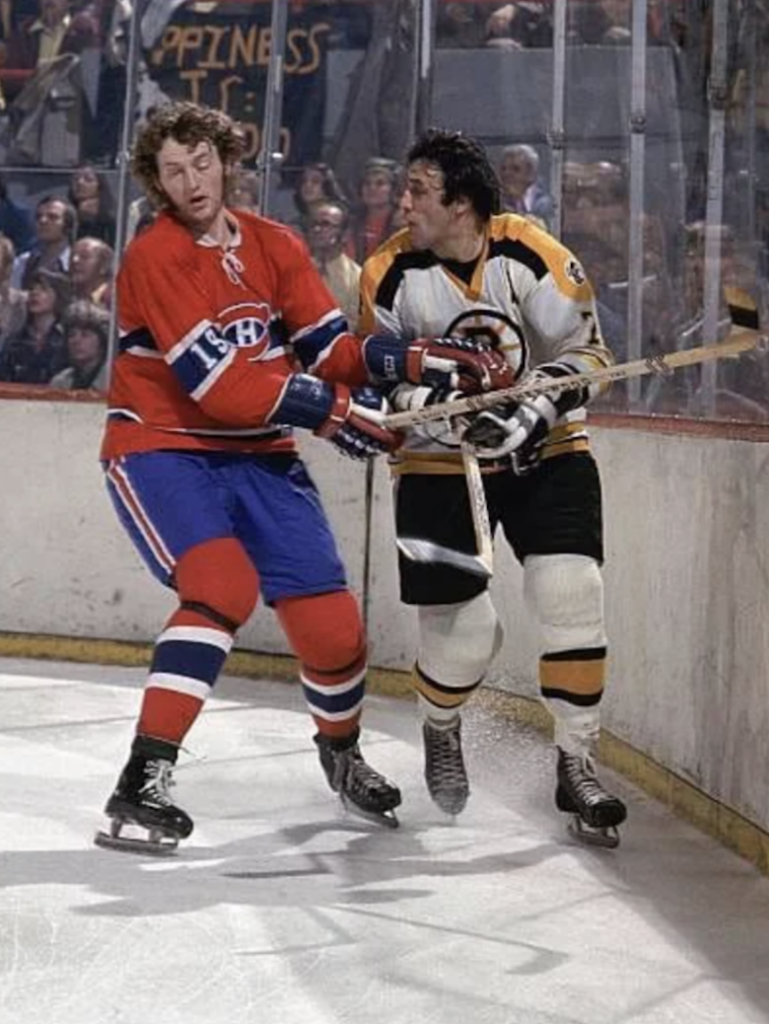 Two ice hockey players in action on the rink. One wears a red and blue uniform, while the other wears a white and yellow uniform. They are near the boards, with a crowd in the background watching the game.