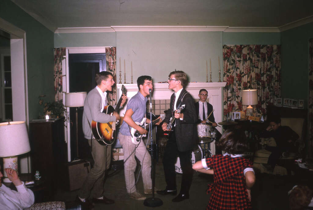 A retro scene of a band performing in a living room. Three members play guitars and sing into microphones, while a drummer plays in the background. A young girl dances in the foreground. The room has vintage decor and floral curtains.