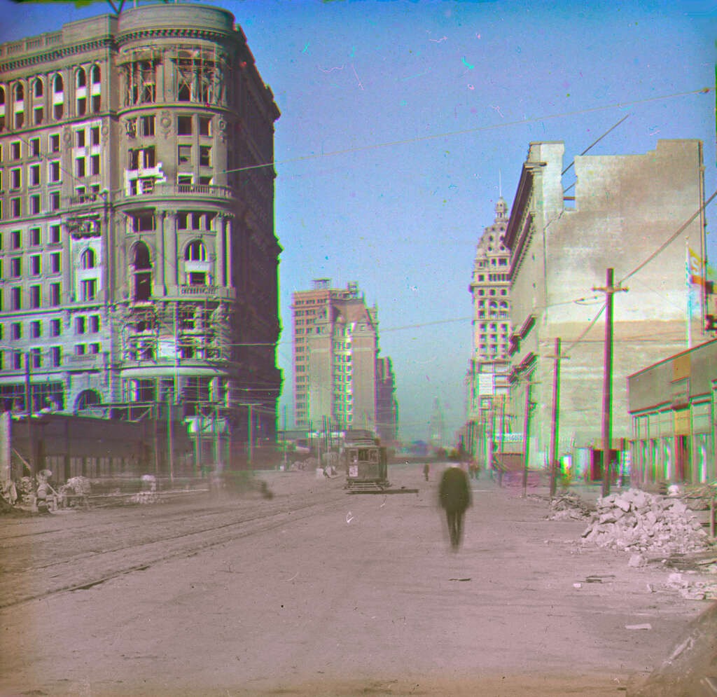 A vintage photograph of a city street features a lone pedestrian walking towards a tram in the distance. On the left, a tall, ornate building stands near another high-rise, while a simpler structure is on the right. Rubble is scattered in the foreground.