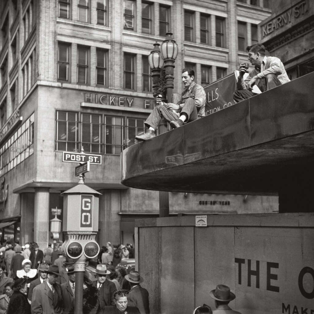 People gathered at a busy intersection near construction workers sitting on a ledge in a city. The scene includes vintage street signs and buildings with large windows. The men on the ledge appear relaxed, watching the crowd below.