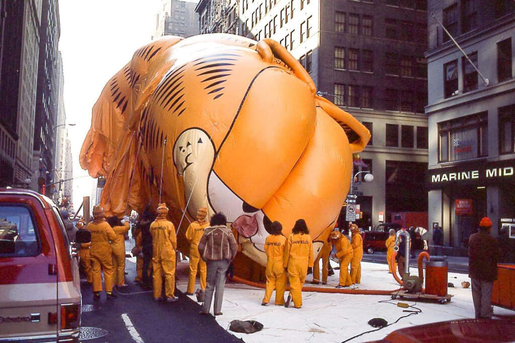 early 80s photos - A large orange inflatable cartoon character is deflating on a city street surrounded by tall buildings. Several people in orange uniforms stand around it, some are holding ropes, and a few vehicles are parked nearby.