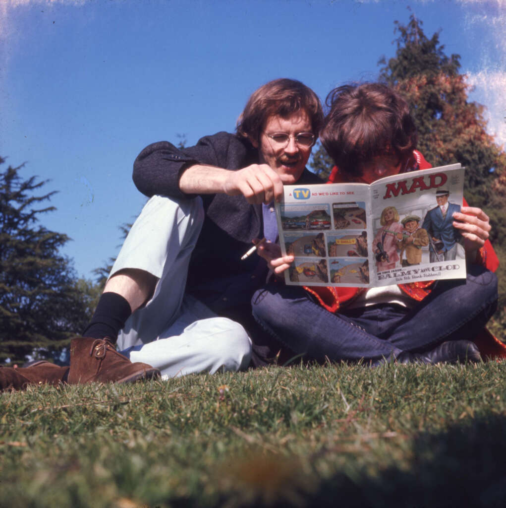 Two people sitting on grass, engrossed in reading a MAD magazine. One is wearing glasses and a dark jacket, while the other wears a red jacket. Clear blue sky and trees in the background.