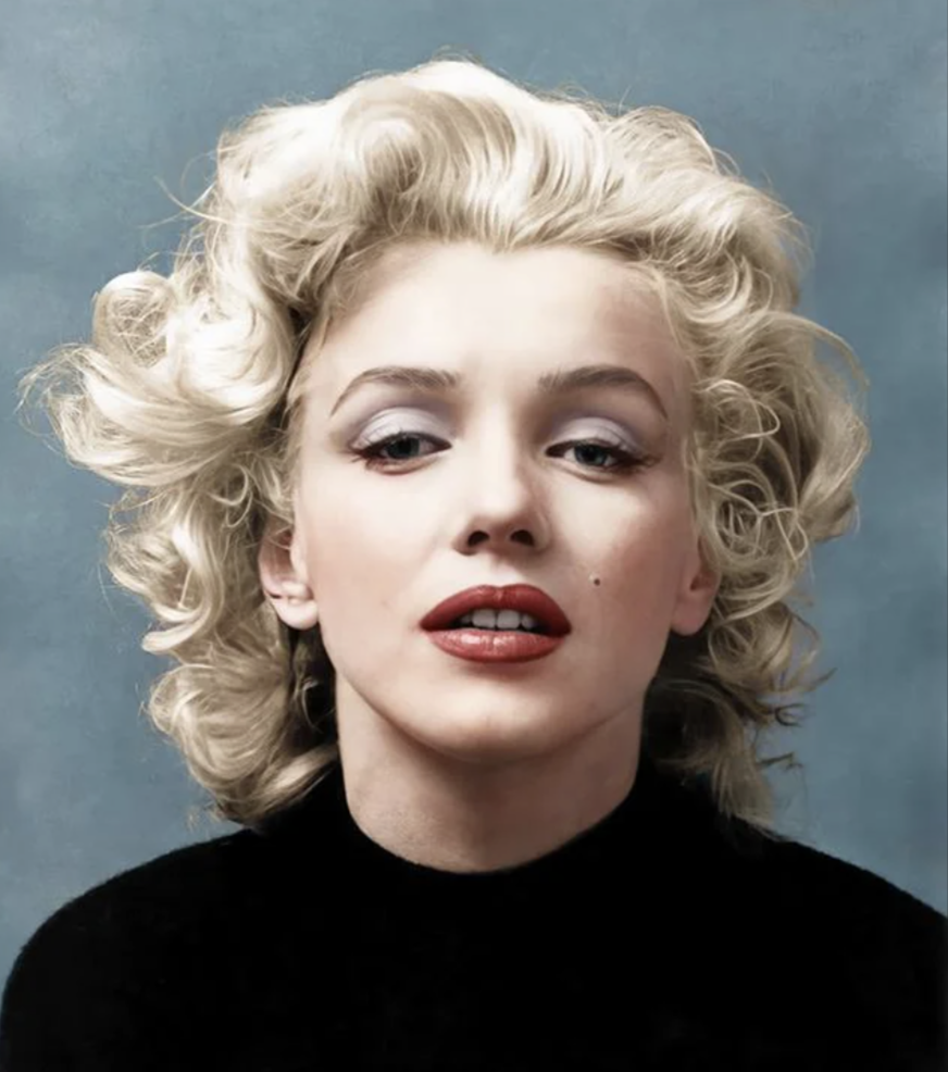 A person with curly blonde hair and red lipstick gazes into the camera against a blue background, wearing a black top.