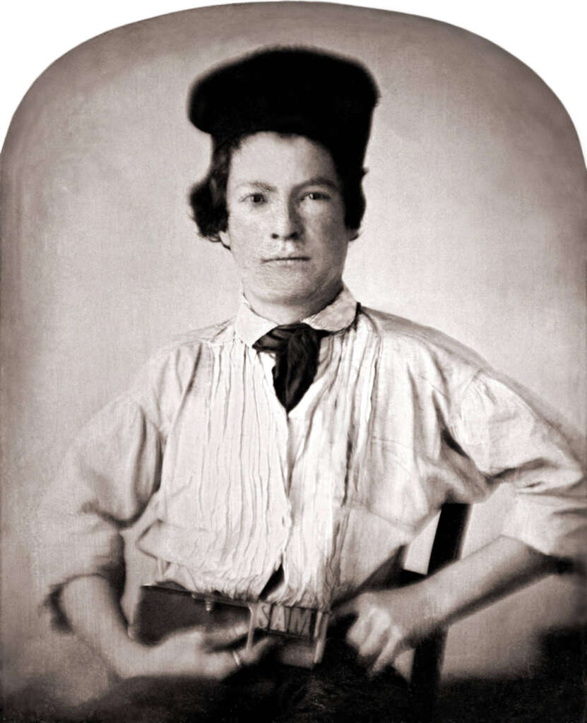 Vintage portrait of a young person in a light shirt with a dark cravat and cap, sitting on a chair. The person has a neutral expression and is wearing a belt with the name "Sam" on the buckle. The image is sepia-toned.