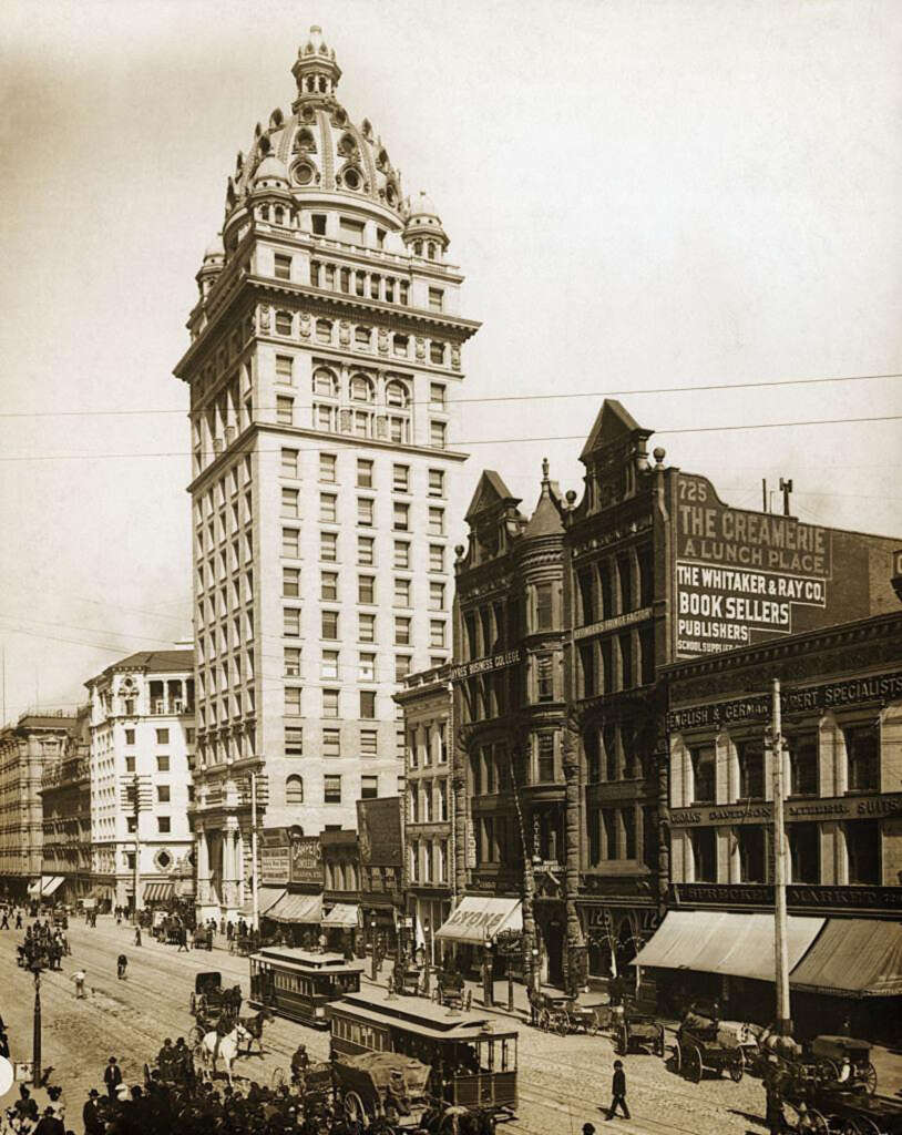 A vintage photo shows a bustling city street with a tall, ornate building, likely from the early 20th century. Horse-drawn carriages and pedestrians fill the street. Storefronts and signs, including one for booksellers, are visible along the road.