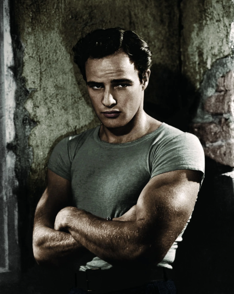 Man with dark hair stands against a textured wall, arms crossed, wearing a light gray t-shirt. His expression is serious. The scene has a vintage, classic look.