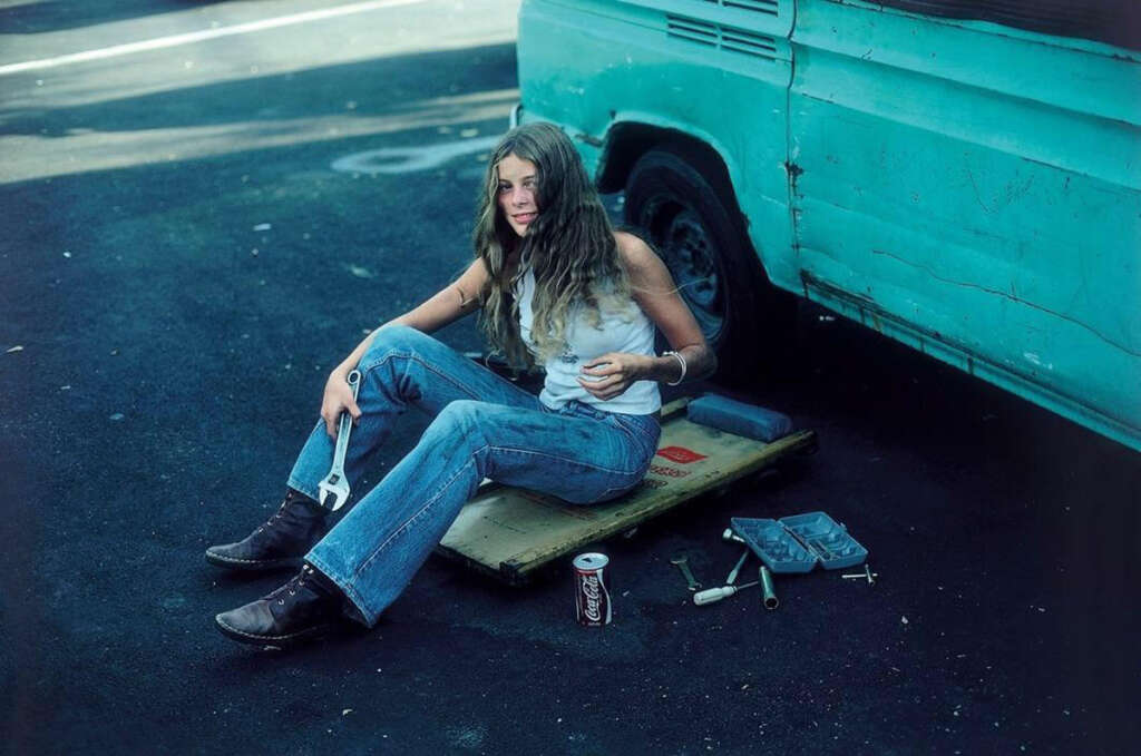 A person with long hair, wearing a tank top and jeans, sits on a wooden platform next to a turquoise van. They are holding a wrench. Nearby are a can and a tray. The setting appears to be a parking lot.