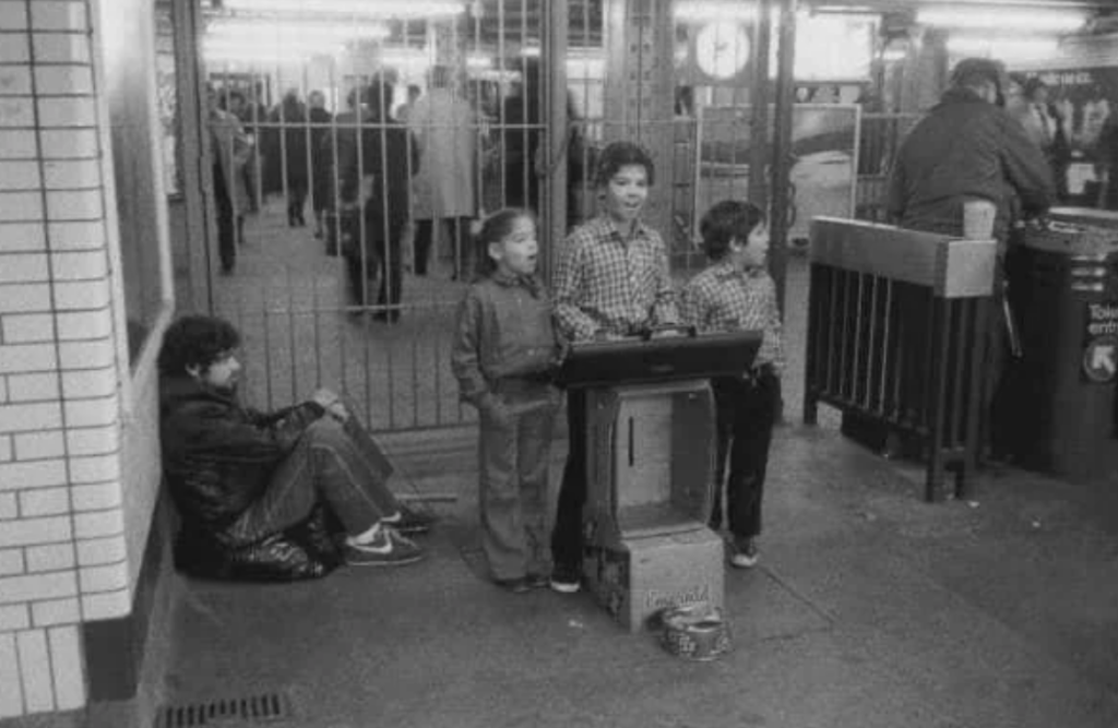 Three children sing next to a keyboard on a subway platform, with one child playing the instrument. A person sits against the wall watching them. A few commuters walk in the background. The setting is dimly lit and appears historical.