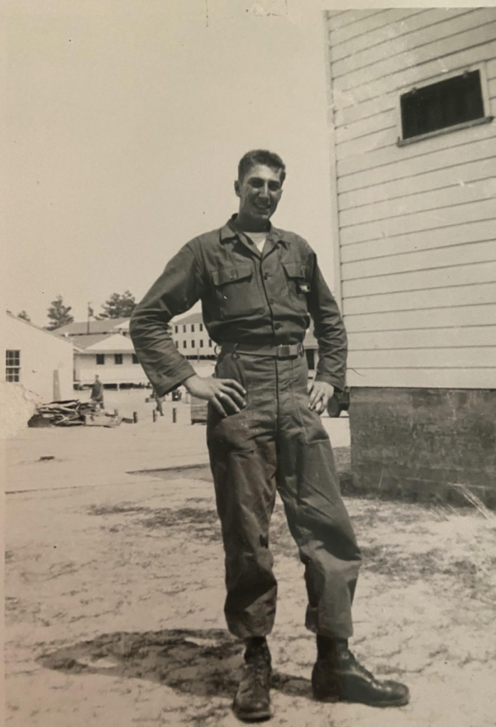 Black and white photo of a man in military uniform standing outside with hands on hips, in front of a building. Background includes another building and a person. The setting appears to be a military base or camp.