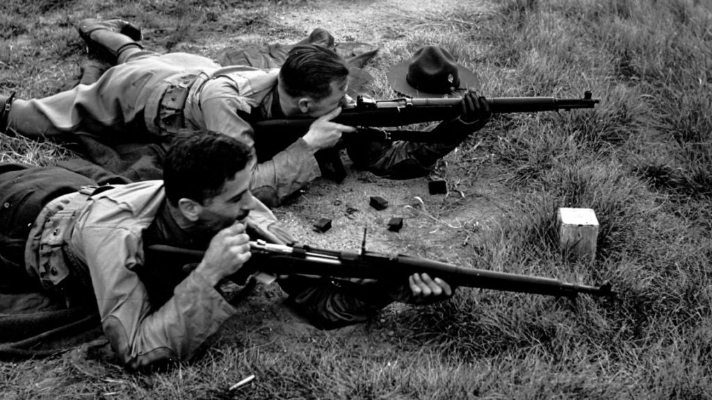 Two soldiers lying prone on grass, aiming rifles. One wears a brimmed hat placed beside him. The image is in black and white, capturing a training or combat scenario with scattered equipment around them.