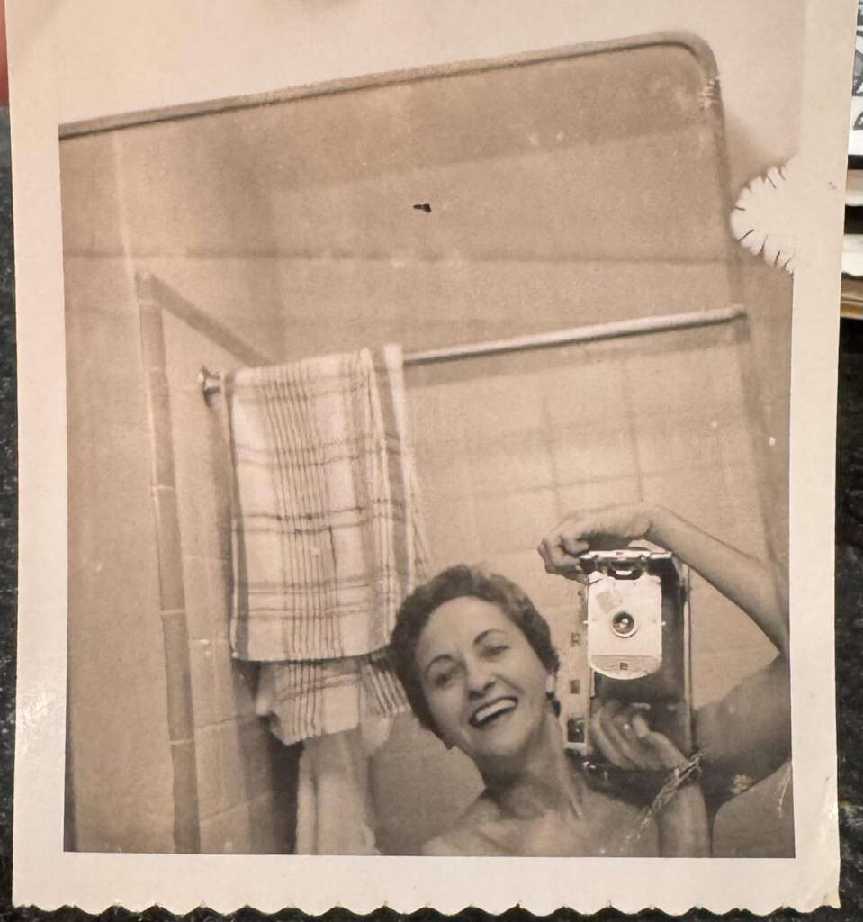 A black-and-white vintage photo of a woman taking a mirror selfie in a bathroom. She is smiling and holding an old-fashioned camera. A plaid towel hangs on the shower door behind her.