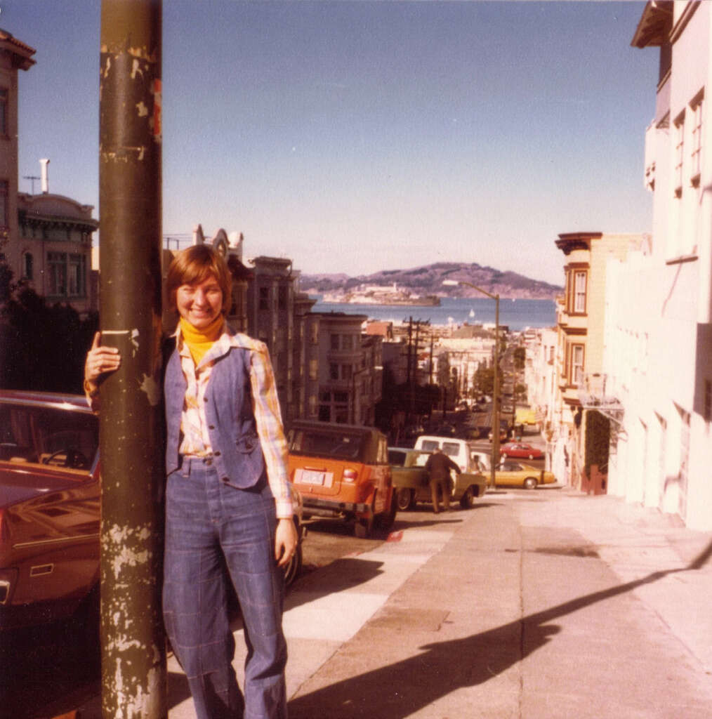 A person in 1970s attire leans against a pole on a steep San Francisco street. Cars are parked along the road, with Alcatraz Island visible in the distance under a clear blue sky.