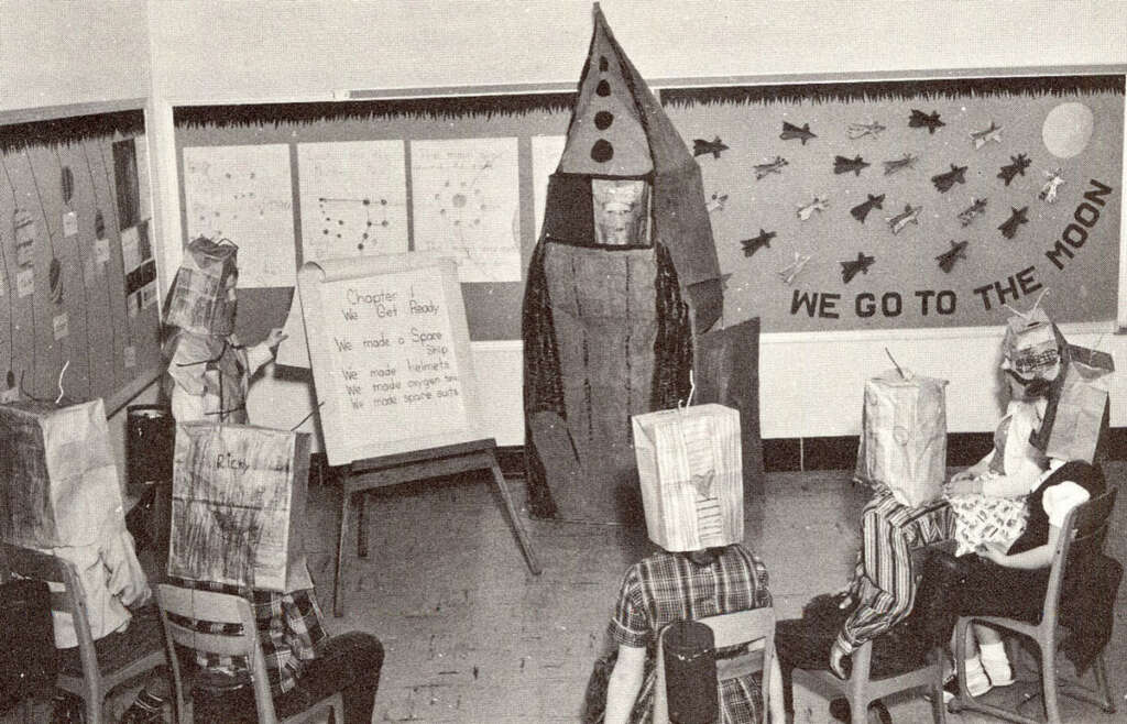 Children in a classroom wearing paper bag helmets sit facing a chalkboard and a person dressed as a rocket. The walls display space-themed art, including a "We Go to the Moon" sign and drawings of rockets and planets.