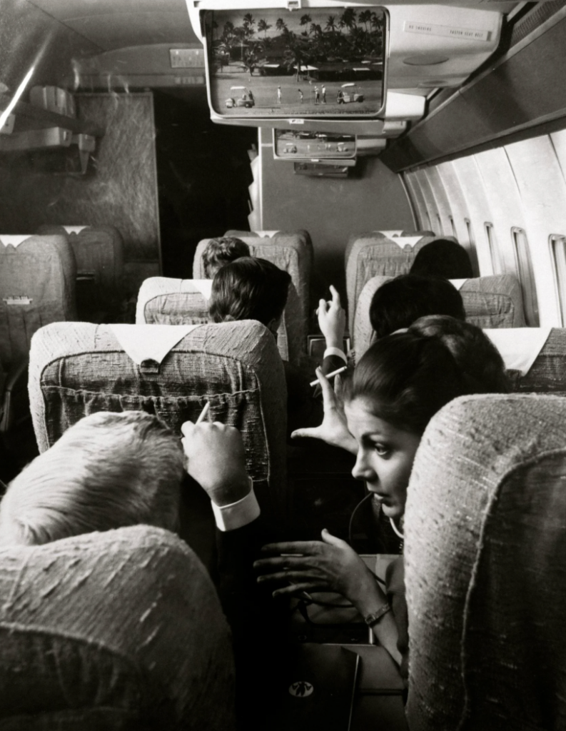 Passengers sitting in an airplane, engaged in conversation. Some are pointing at a screen overhead displaying a tropical scene. The image is in black and white, giving a vintage feel to the airplane's interior.