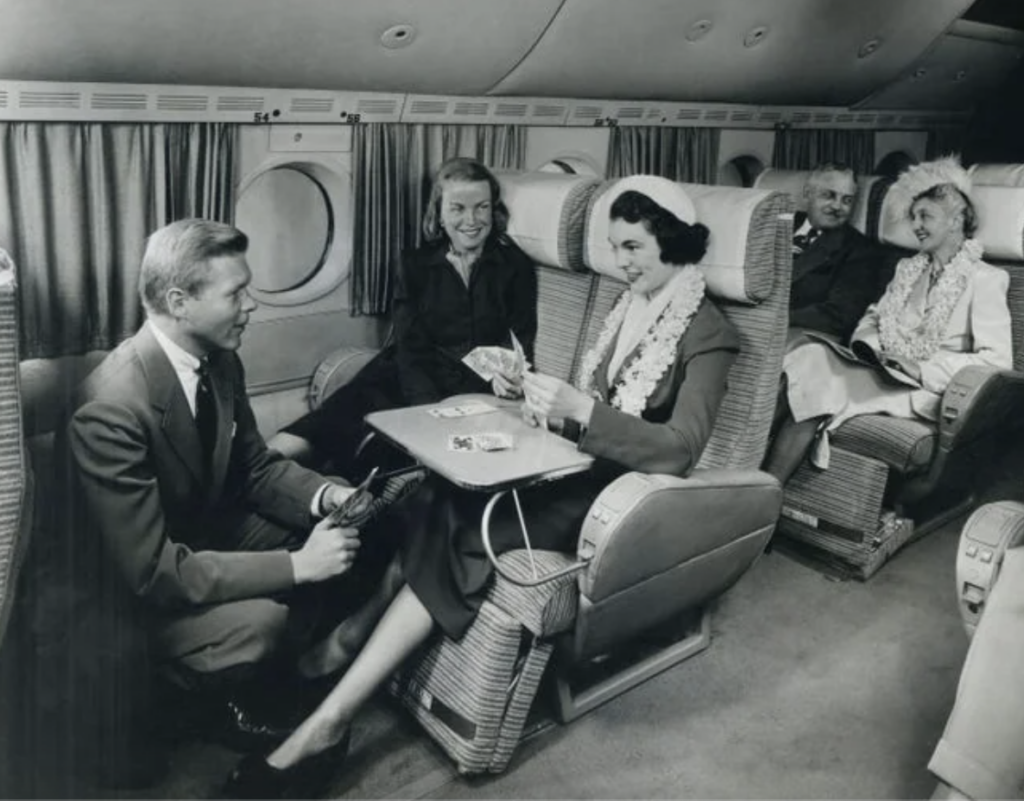 Passengers in vintage airline seats play cards and read. A man is crouched by the table with a hand of cards. Curtains cover the windows, and two women in hats are seated at the back, with a man reading a magazine. The setting appears elegant and classic.