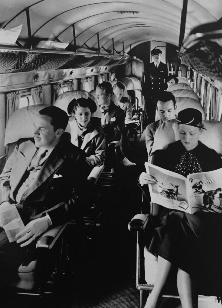 Black and white photo of six passengers seated in an airplane cabin, dressed in mid-20th century attire. The passengers are reading and talking, with a flight attendant standing at the back. The cabin has vintage-style seats and overhead storage.