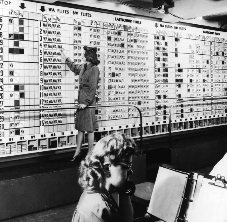 A black-and-white photo of a woman standing on a platform pointing at a large board with numerous flight schedules and codes. Another woman sits at a desk below, using early aviation communication equipment.