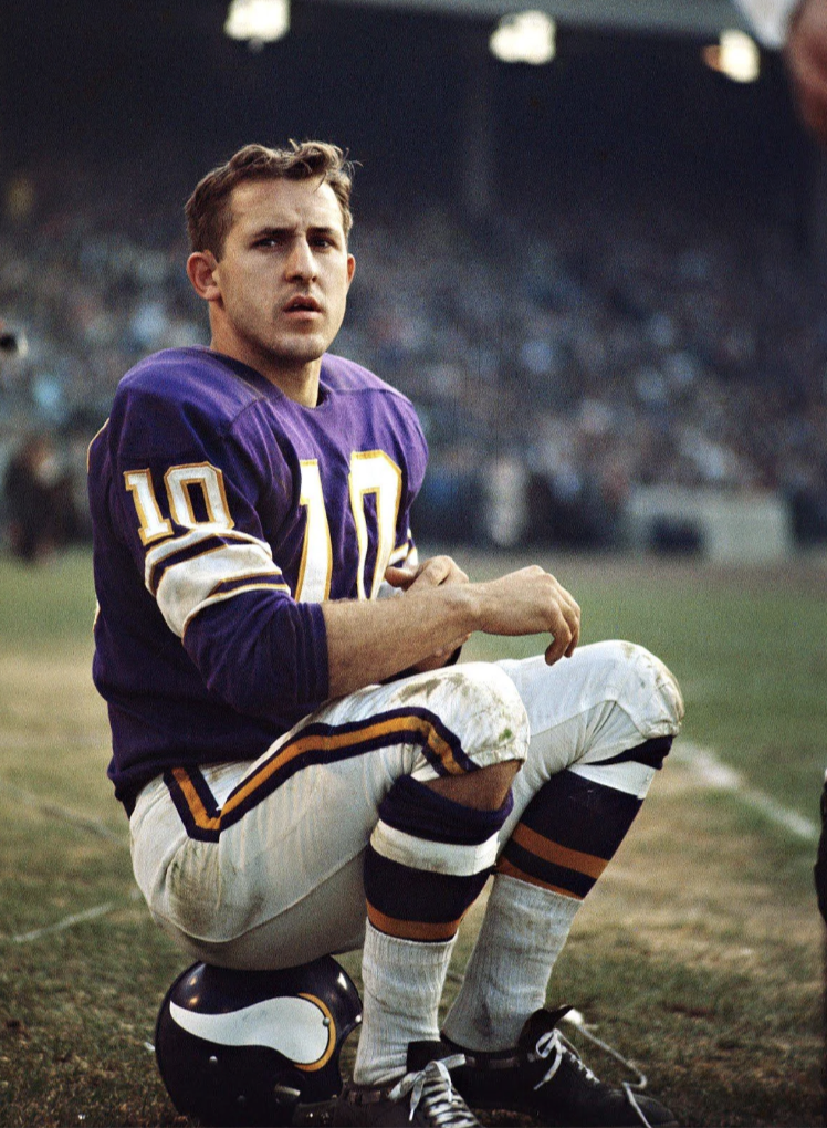 Football player in a purple and white uniform with the number 10 sits on a helmet on the sidelines of a field. The background shows a stadium with blurred spectators.