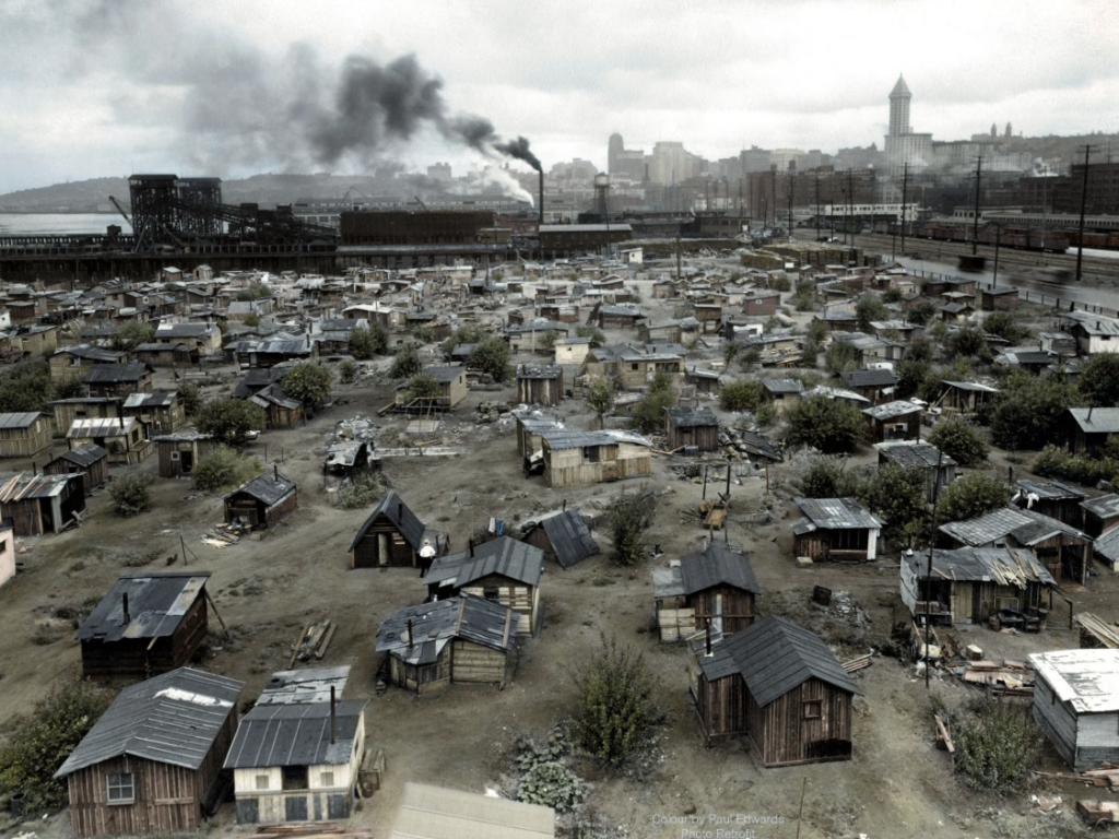 A depression-era shantytown with numerous makeshift homes is set amid a barren landscape. Smoke rises from a factory in the background, with a city skyline visible under a cloudy sky.