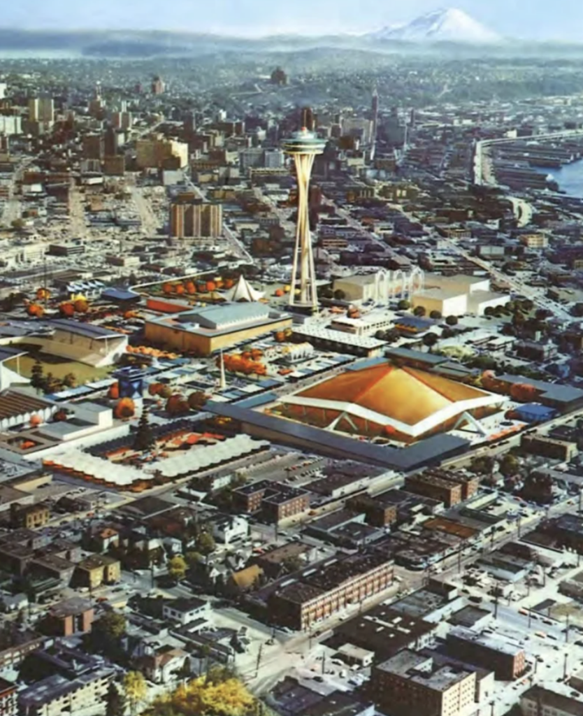 Aerial view of Seattle's cityscape featuring the Space Needle at the center. Buildings and streets surround the area, with mountains visible in the distant background. The image captures a clear, sunny day.