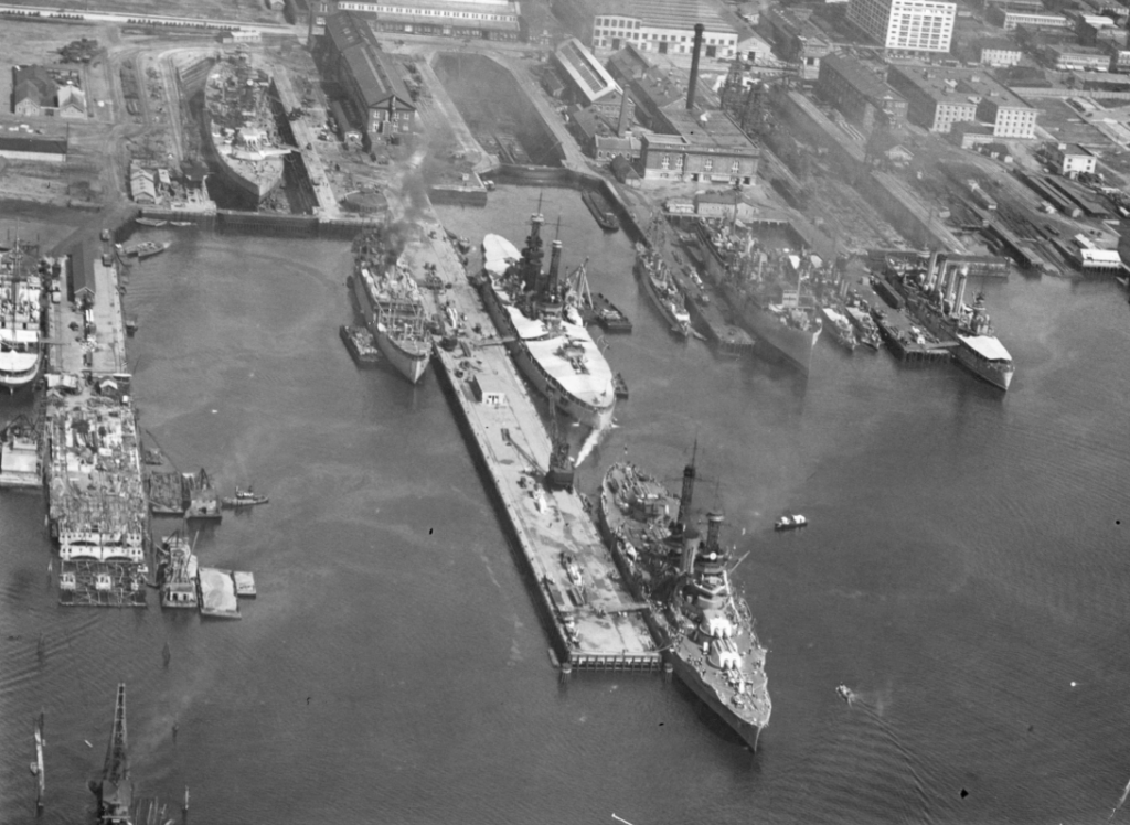 Aerial view of a shipyard with several large naval vessels docked. The area is bustling with ships under construction or repair, surrounded by industrial buildings and cranes.