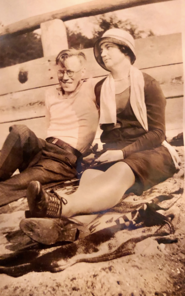 Vintage photo of a man and woman sitting on a sandy beach. The man is wearing glasses and a light-colored shirt, while the woman is in a dress and cloche hat. Both are relaxed, leaning against a wooden structure, with a towel on the sand.