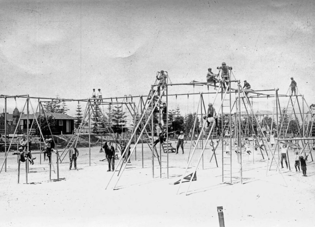 Children playing on large metal playground equipment at a park. Some are climbing ladders, while others use swings. The scene is lively with many kids actively engaged in play. Trees and buildings are visible in the background. The image is in black and white.