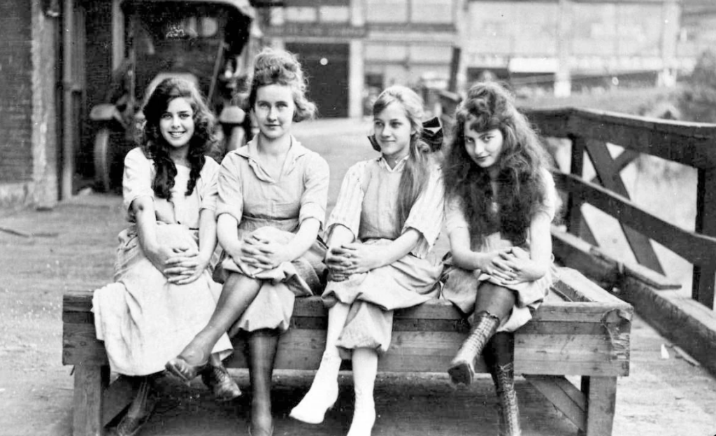 Four young women sit on a wooden platform, smiling for the camera. They are dressed in early 20th-century attire, with long skirts and blouses. An old vehicle is partially visible in the background on a cobblestone street.