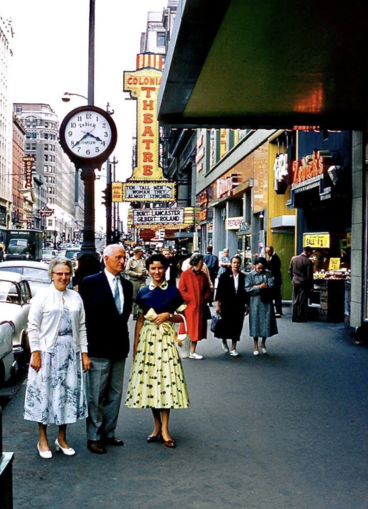 Street scene from the 1950s showing people walking on a city sidewalk. A man and two women pose for a photo. Theater marquees, a clock, and cars are visible. The women wear dresses, and the man is in a suit. Buildings line the street.