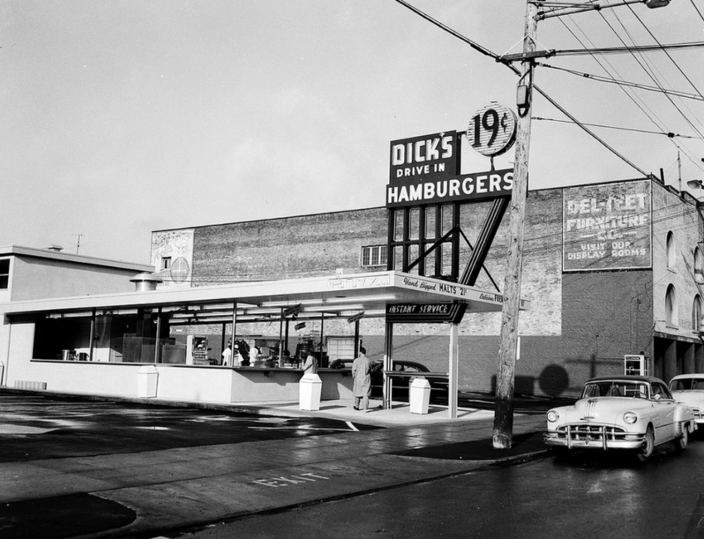 Black and white photo of a vintage drive-in restaurant named "DICK'S Drive In Hamburgers" with a large sign showing 19-cent burgers. A classic car is parked in front, and a few people are visible inside the open-air counter service area.