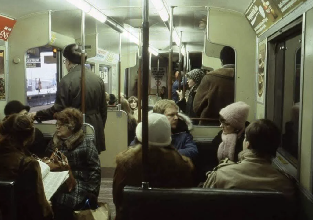 People in winter clothing sit and stand inside a vintage subway car. Some are engaged in conversation, while others read or look out the windows. The interior is dimly lit, with ads above the seats. An outside platform is visible through the windows.