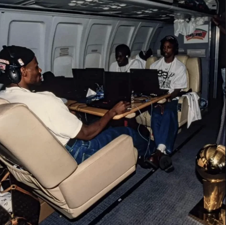 Three people are sitting and using laptops on a private jet, with one wearing headphones. The cabin has leather seats and a table. A golden trophy sits near them on the floor. They appear relaxed, engaged in their devices.