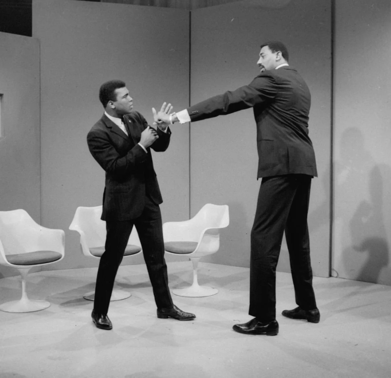 Two men in formal suits are playfully posing on a stage; one is reaching his long arm towards the other, who assumes a boxing stance. Three modern white chairs are in the background.