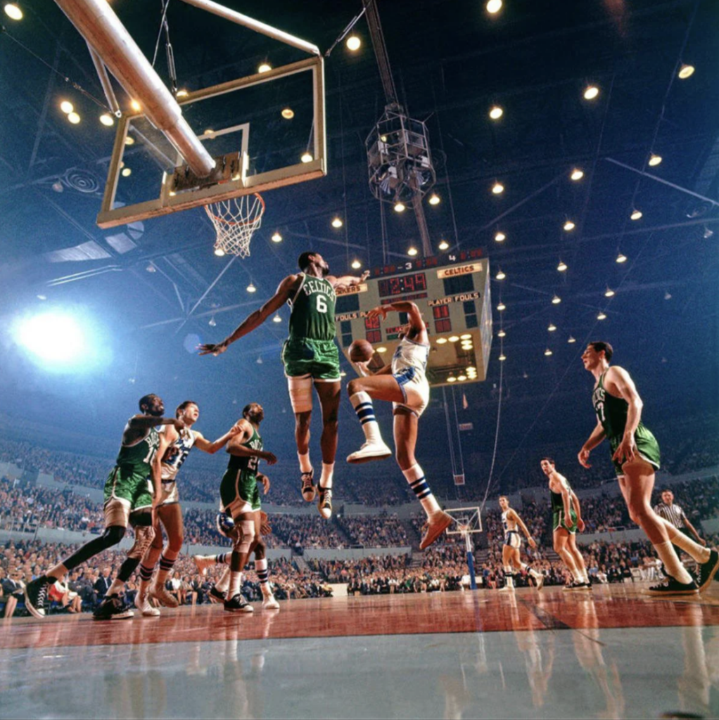 Basketball game in a packed indoor arena. Players, wearing green and white uniforms, are mid-action near the hoop. One player jumps high, attempting a block or dunk, while others prepare to rebound or defend. Bright lights illuminate the scene.