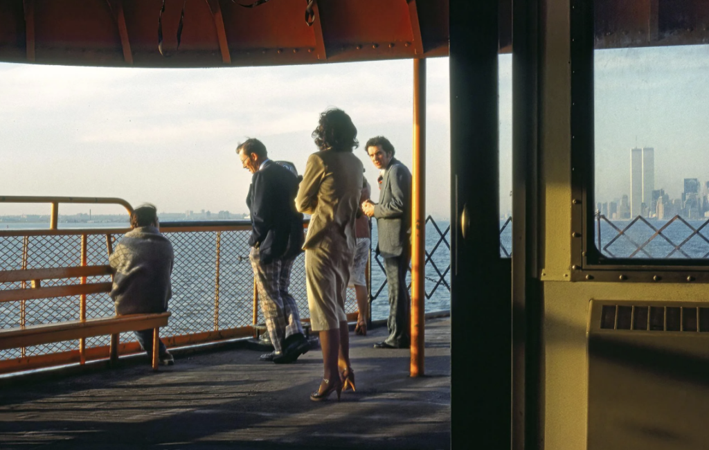 People are standing on a ferry deck overlooking the water. A woman in a light-colored dress gazes outward, while others appear relaxed. The skyline, including the Twin Towers, is visible in the background under a partly cloudy sky.