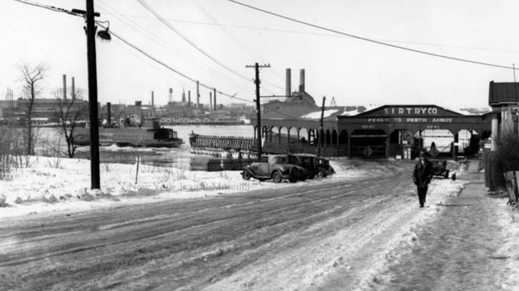 A snowy road leads to a ferry terminal with a sign reading "PERTH AMBOY." There are vintage cars parked along the side, a person walking, and industrial buildings in the background. The scene is overcast and wintry.