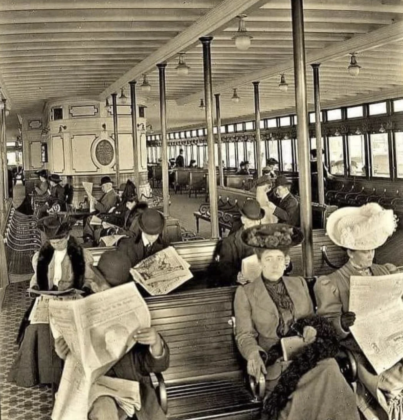 A vintage photograph shows passengers on a historic train or tram. Several men and women, dressed in early 20th-century attire, are seated on wooden benches. Some read newspapers, and one woman wears a hat with a large feather. The interior is ornate.