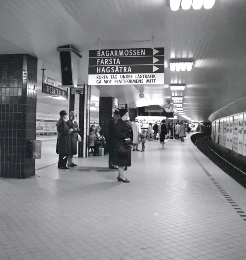 Black-and-white photo of a subway station platform with several people standing and waiting. Overhead signs point towards Bagarmossen, Farsta, and Hagsätra. The tunnel curves to the right, and the station is tiled with overhead lighting.