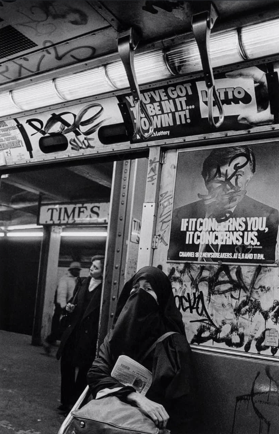 A person in a niqab stands inside a graffiti-covered subway train holding a newspaper. A poster on the wall reads, "IF IT CONCERNS YOU, IT CONCERNS US." Another person is visible outside the train. Posters and graffiti cover the surroundings.