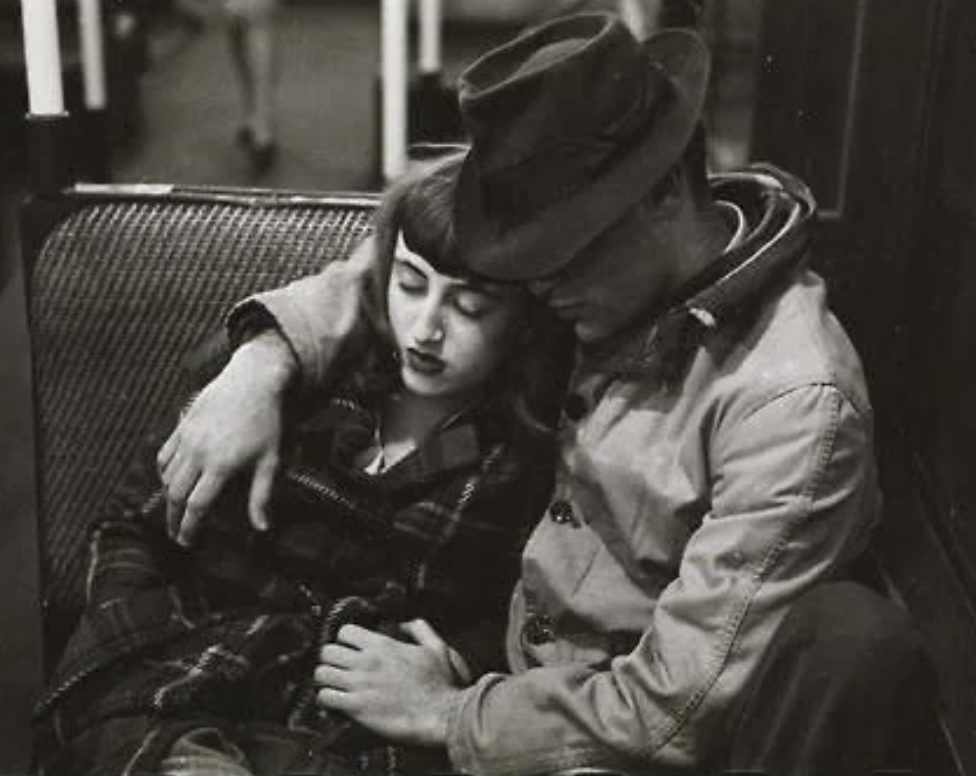 A black and white photo of a couple asleep on public transport. The man wears a hat and coat, resting his head on hers, while the woman has long hair and a checkered coat, leaning on his shoulder. They sit closely, embracing each other.