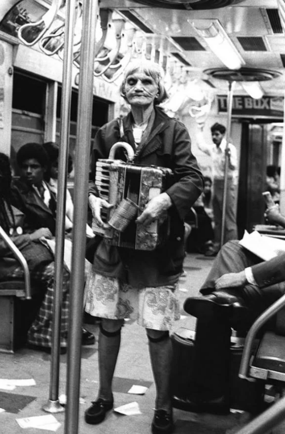 An elderly woman stands in a subway car, playing an accordion. Passengers are seated on either side, some reading newspapers. The floor is scattered with paper. The scene is black and white, lending a vintage feel to the urban setting.